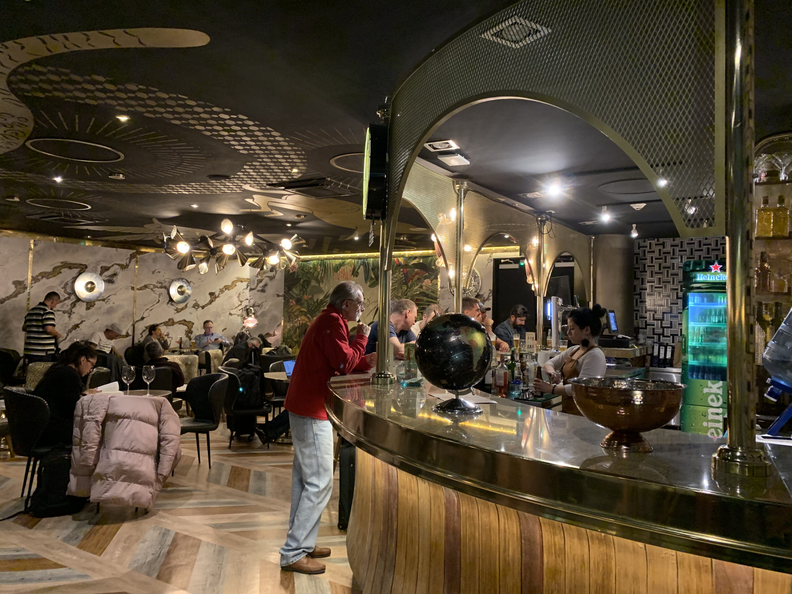 Geschwungene Bar mit Messingverkleidung, Globus-Deko und Heineken-Kühlschrank, Gäste im Hintergrund.