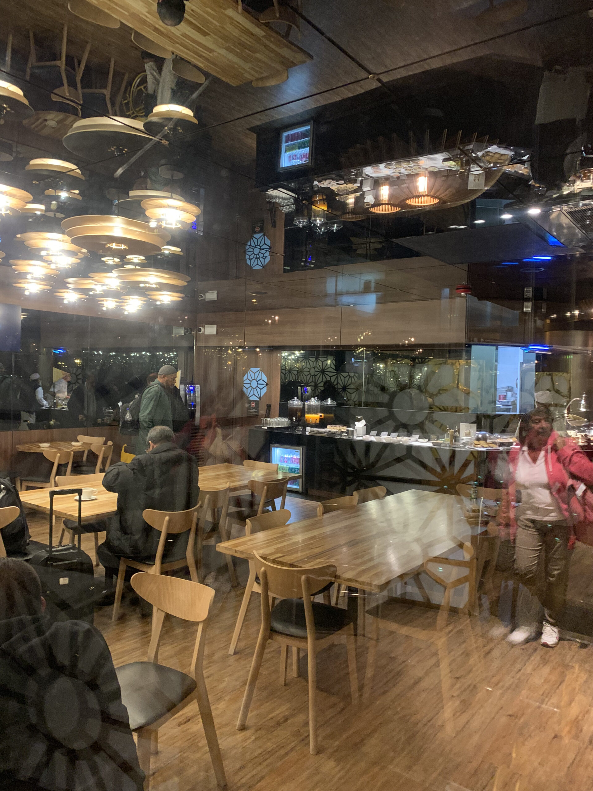 Blick durch eine Glasscheibe in die Lounge mit Holztischen, Stühlen und runden Hängelampen an der Decke.