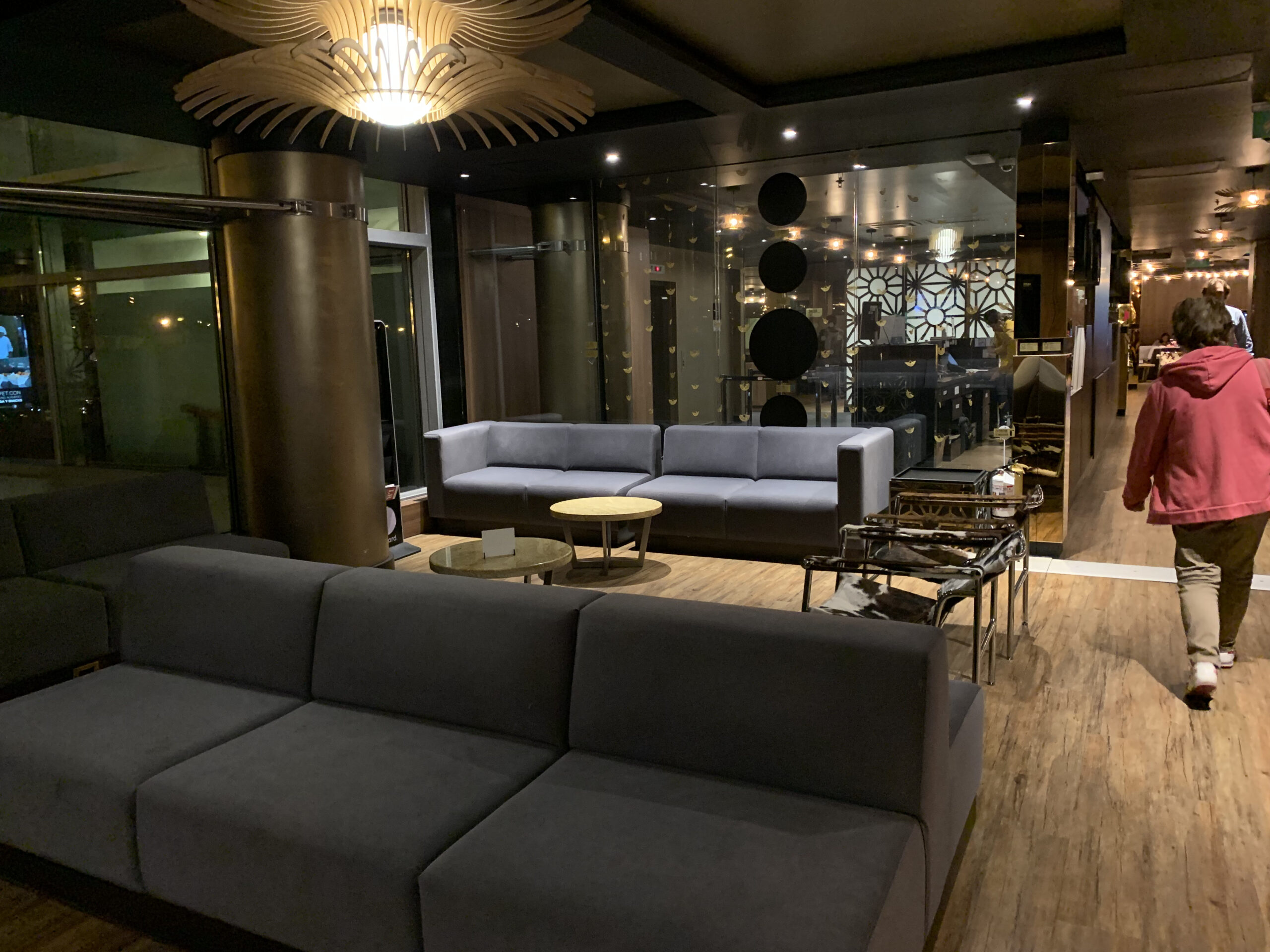 Hotellobby-artiger Loungebereich mit grauen Sofas, Holzboden und dekorativer Deckenleuchte, eine Person läuft vorbei.