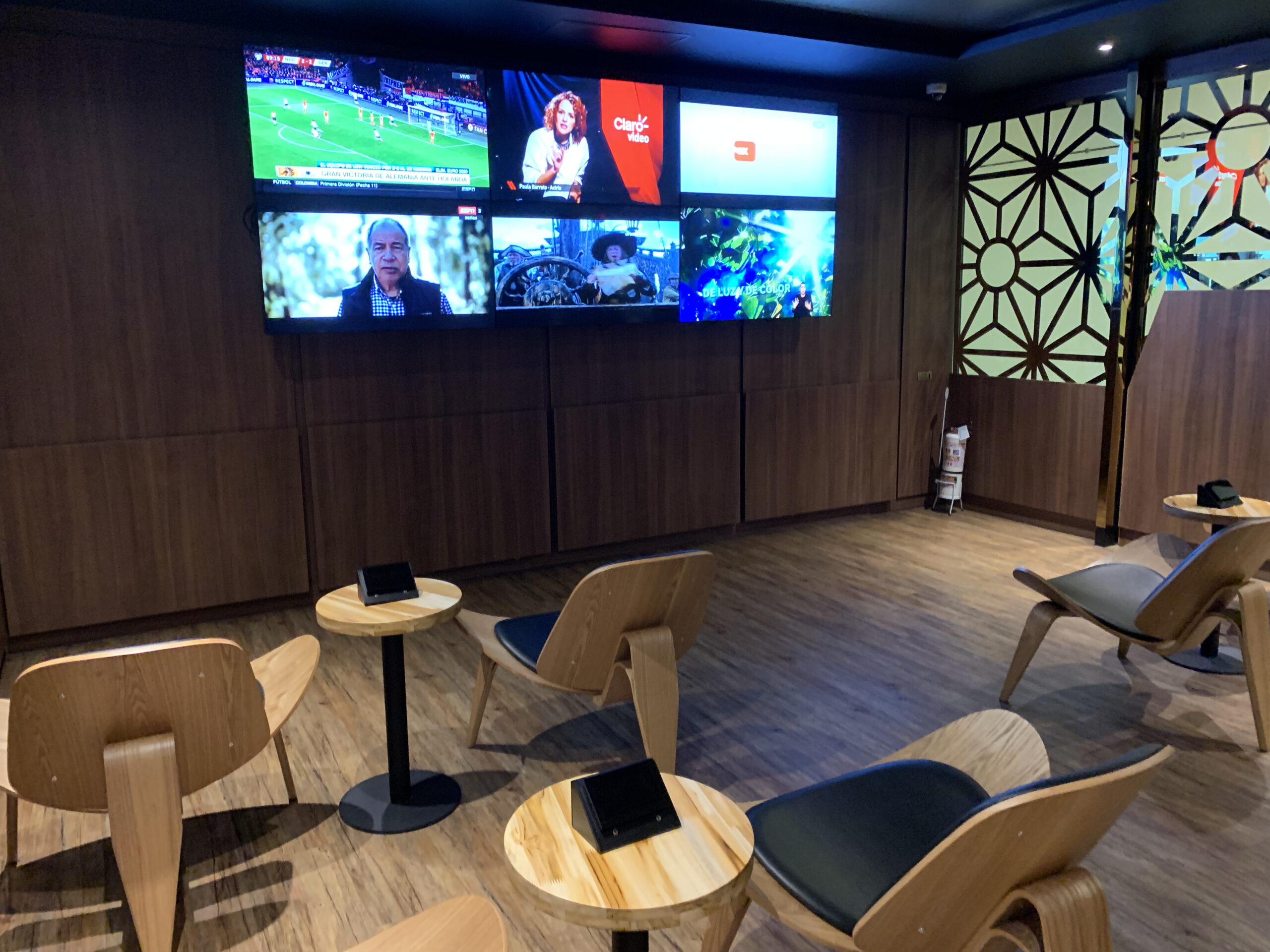 Loungebereich mit sechs Wandbildschirmen, Holzstühlen mit dunklen Sitzpolstern und runden Stehtischen.