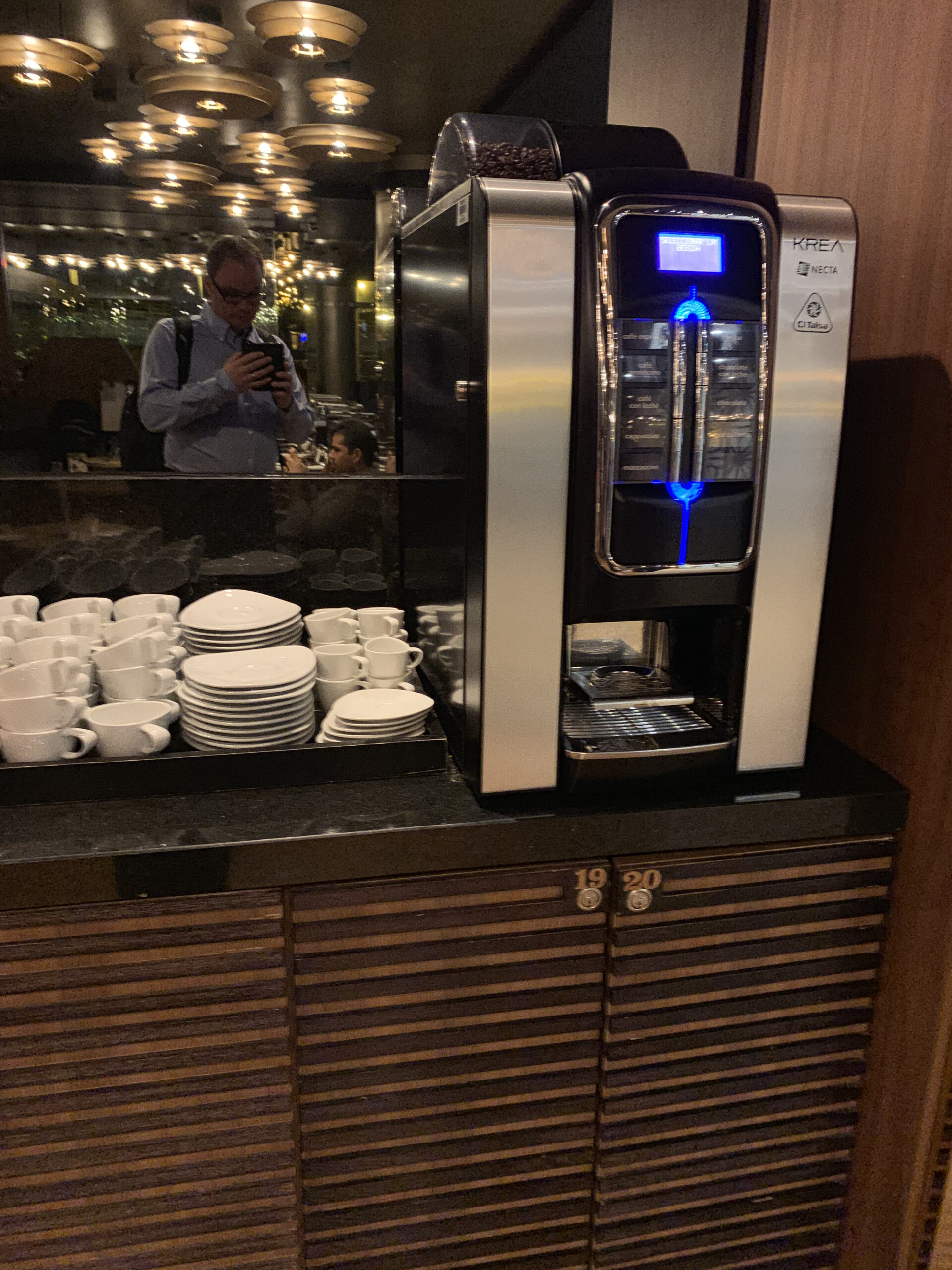 Necta Krea Kaffeevollautomat auf dunkler Theke, daneben Regal mit weißen Tassen und Untertassen.