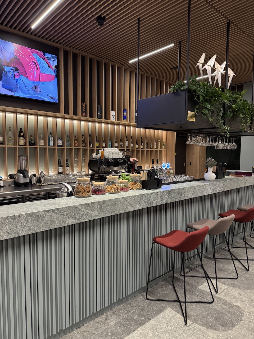 Hotelbar mit grauer Marmortheke, Holzregalwand mit Spirituosen, Espressomaschine und roten Barhockern.