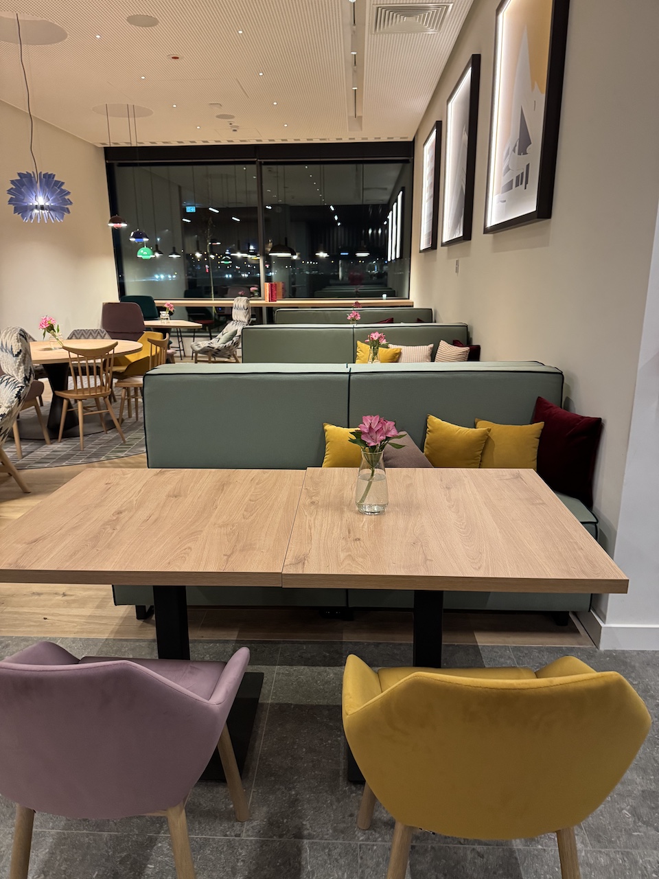 Restaurantbereich mit grünen Sitzbänken, bunten Kissen und Holztischen, im Hintergrund große Fensterfront.