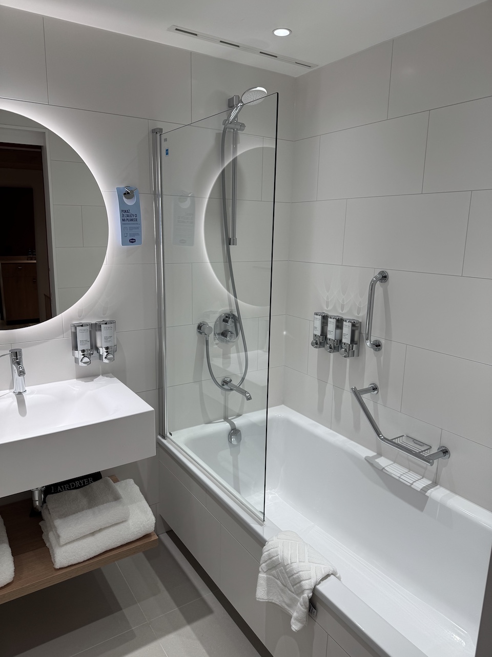 Weißes Hotelbadezimmer mit Badewanne, Duschaufsatz, Glastrennwand, Waschbecken und rundem Spiegel.