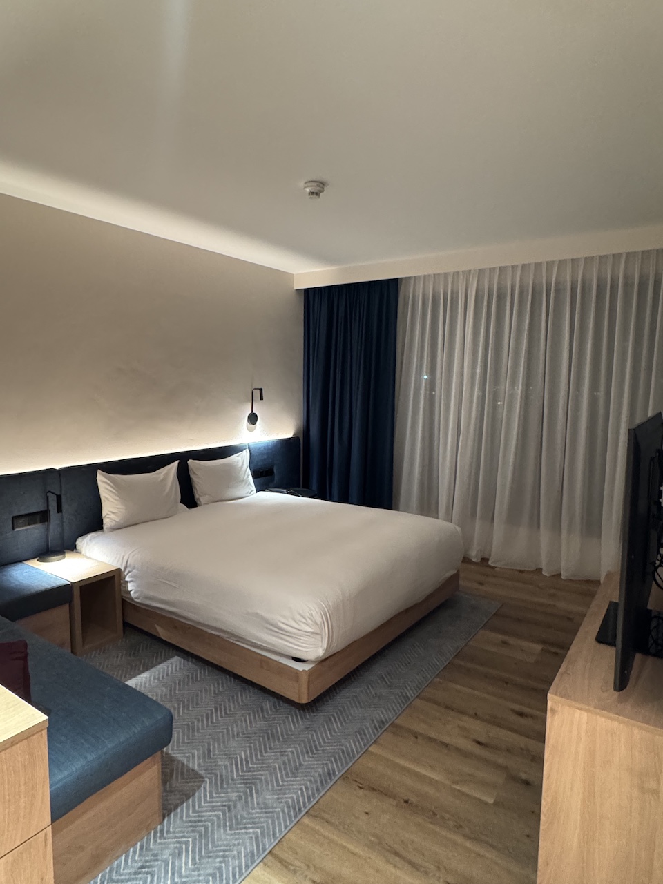 Hotelzimmer mit Doppelbett, dunklem Kopfteil, Holzboden und bodenlangen Vorhängen am Fenster.