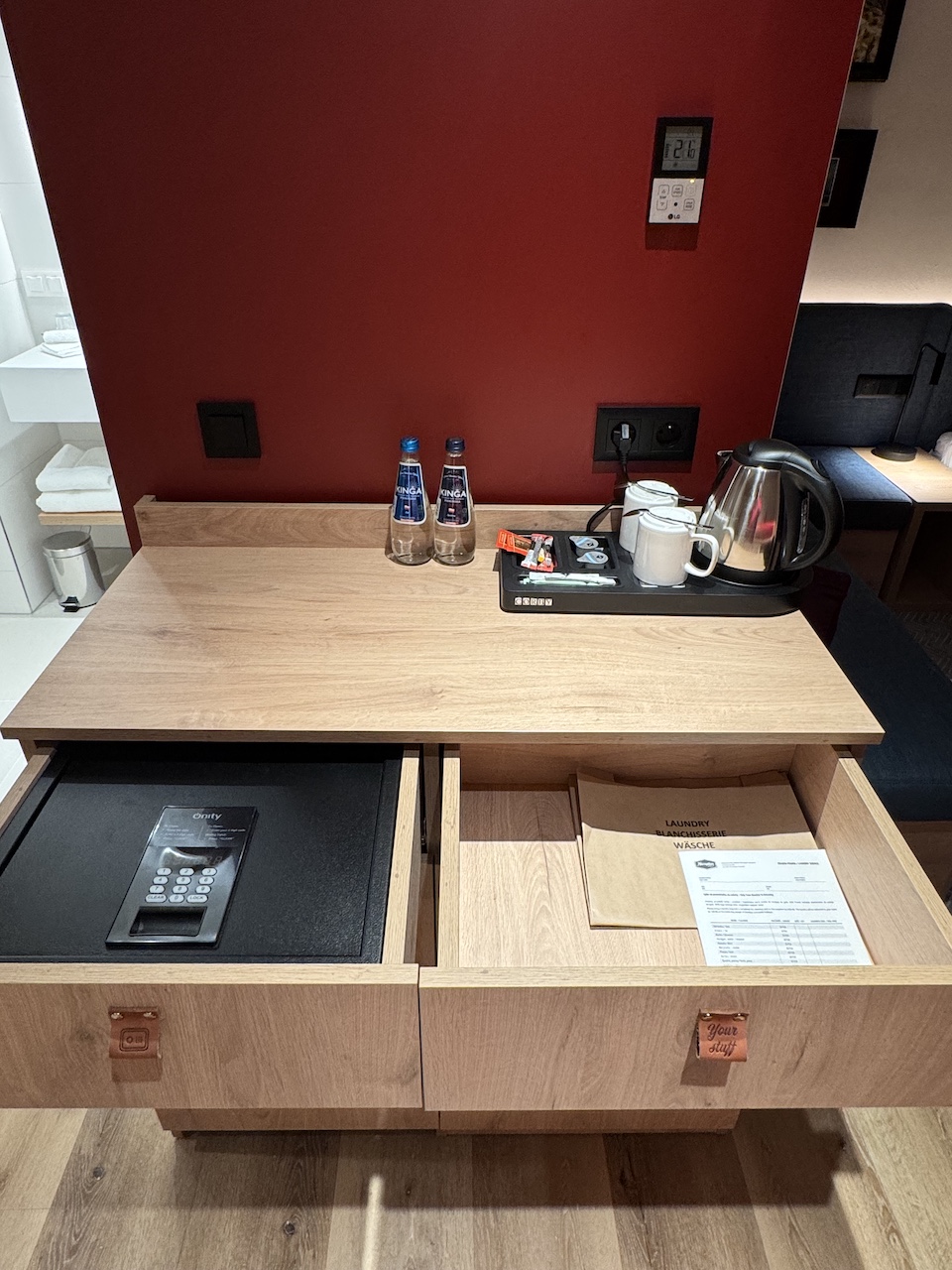 Holzkommode mit geöffneten Schubladen: links Tresor, rechts Wäscheservice-Unterlagen. Oben Wasserkocher und Mineralwasser.