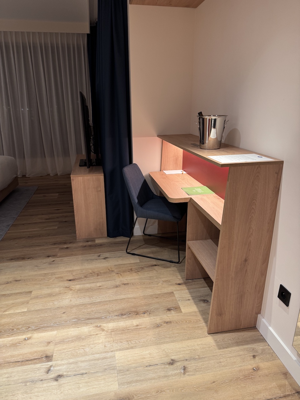 Schreibtisch aus hellem Holz mit grauem Stuhl, Sektkübler und Ablage im Hotelzimmer.