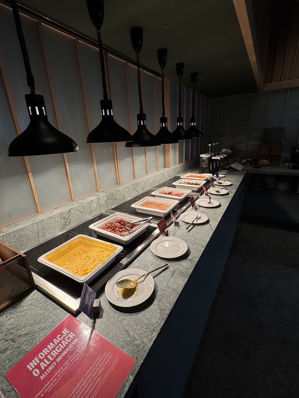 Warme Frühstücksstation mit Rührei, Speck, Würstchen und weiteren Speisen unter schwarzen Wärmelampen.