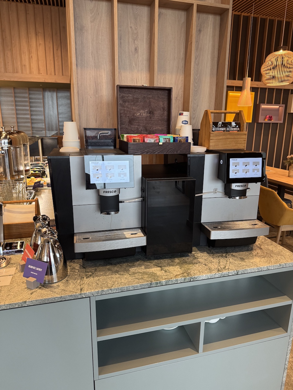 Zwei Fresco-Kaffeevollautomaten auf einer Granittheke mit Teeauswahl, Pappbechern und Thermoskannen.