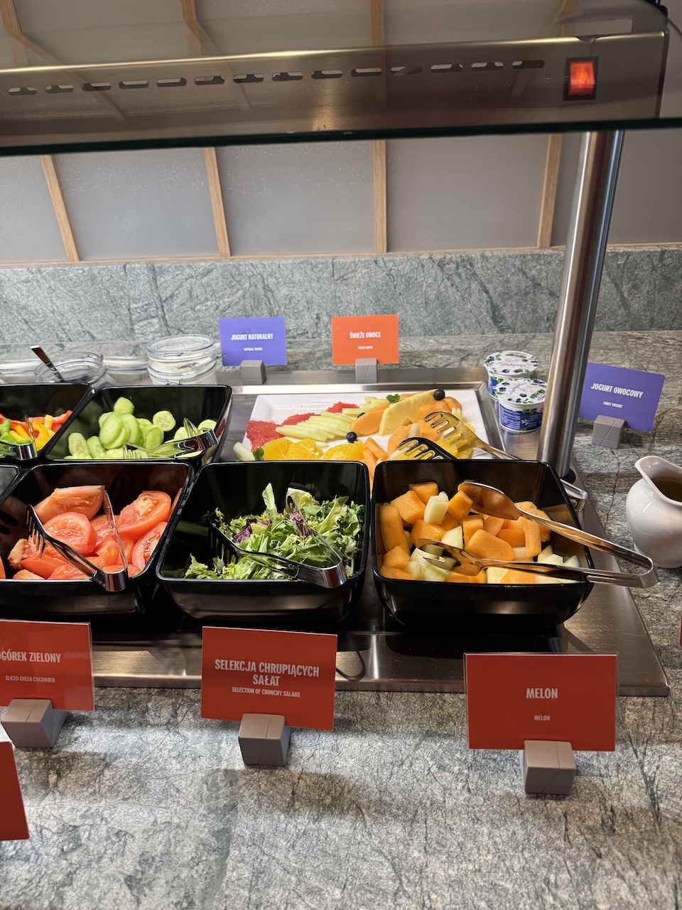 Frühstücksbuffet mit Schüsseln für Gurken, Tomaten, Blattsalat und Melone sowie frische Früchte und Joghurt.