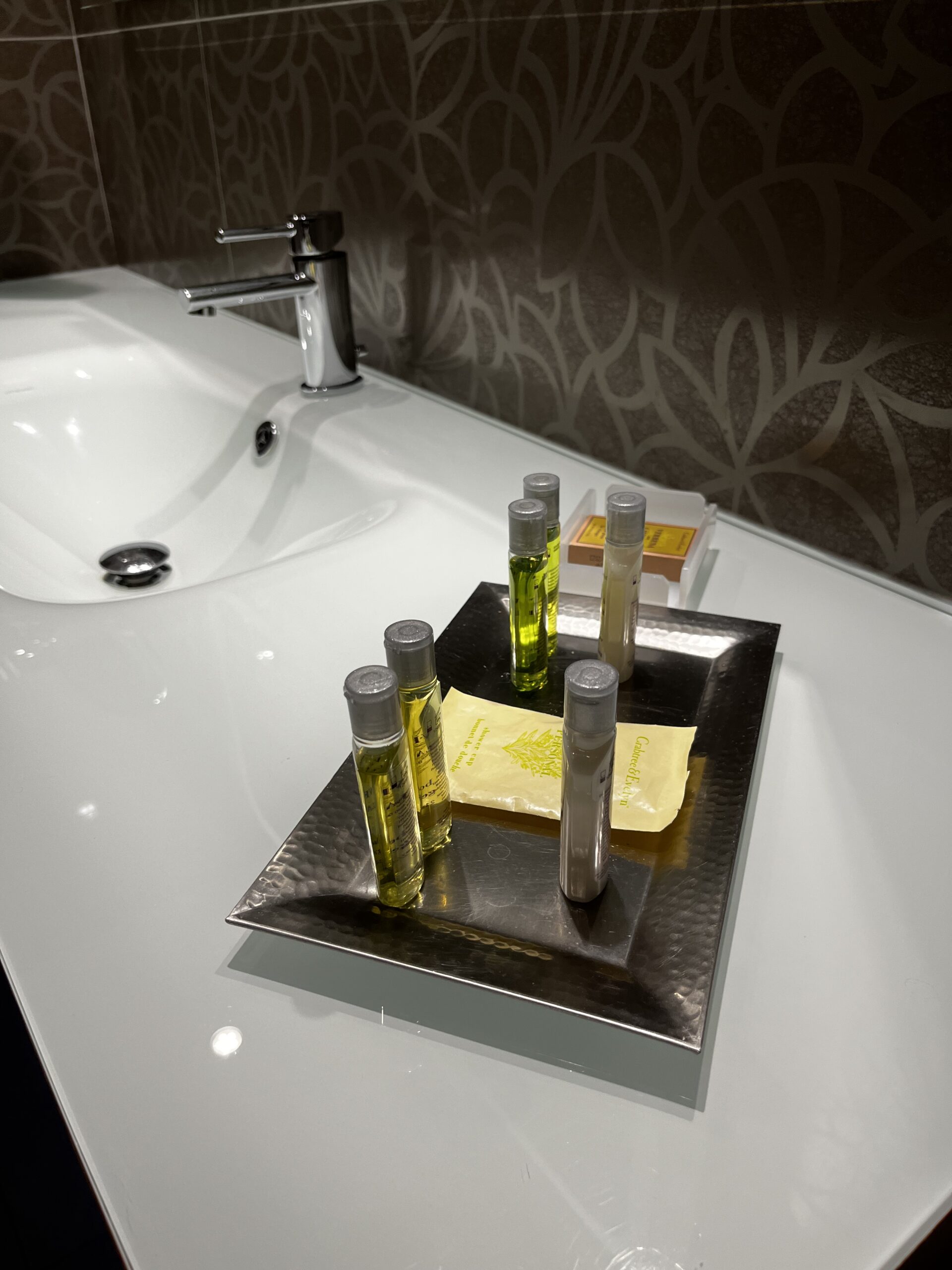 Silbernes Tablett mit mehreren kleinen Shampoo- und Pflegeflaschen sowie Seifenpäckchen auf weißem Waschtisch.