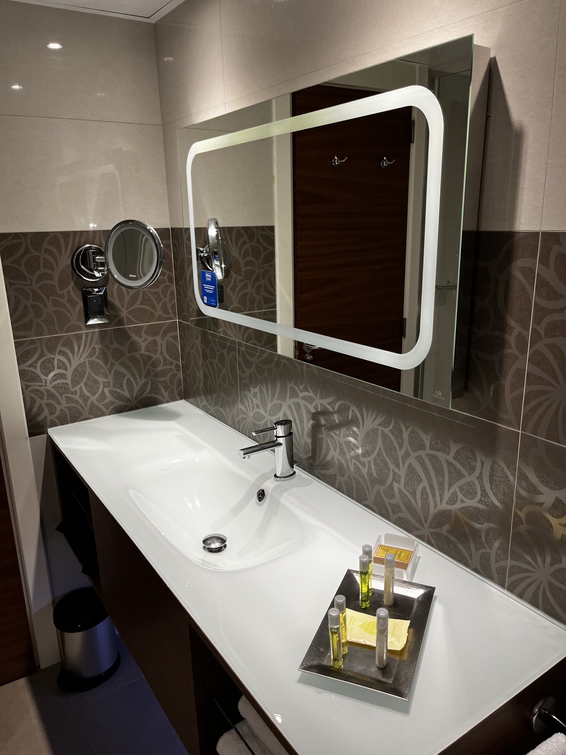 Badezimmer mit weißem Waschtisch, Chromarmatur, beleuchtetem Spiegel und Kosmetikartikeln auf Tablett.