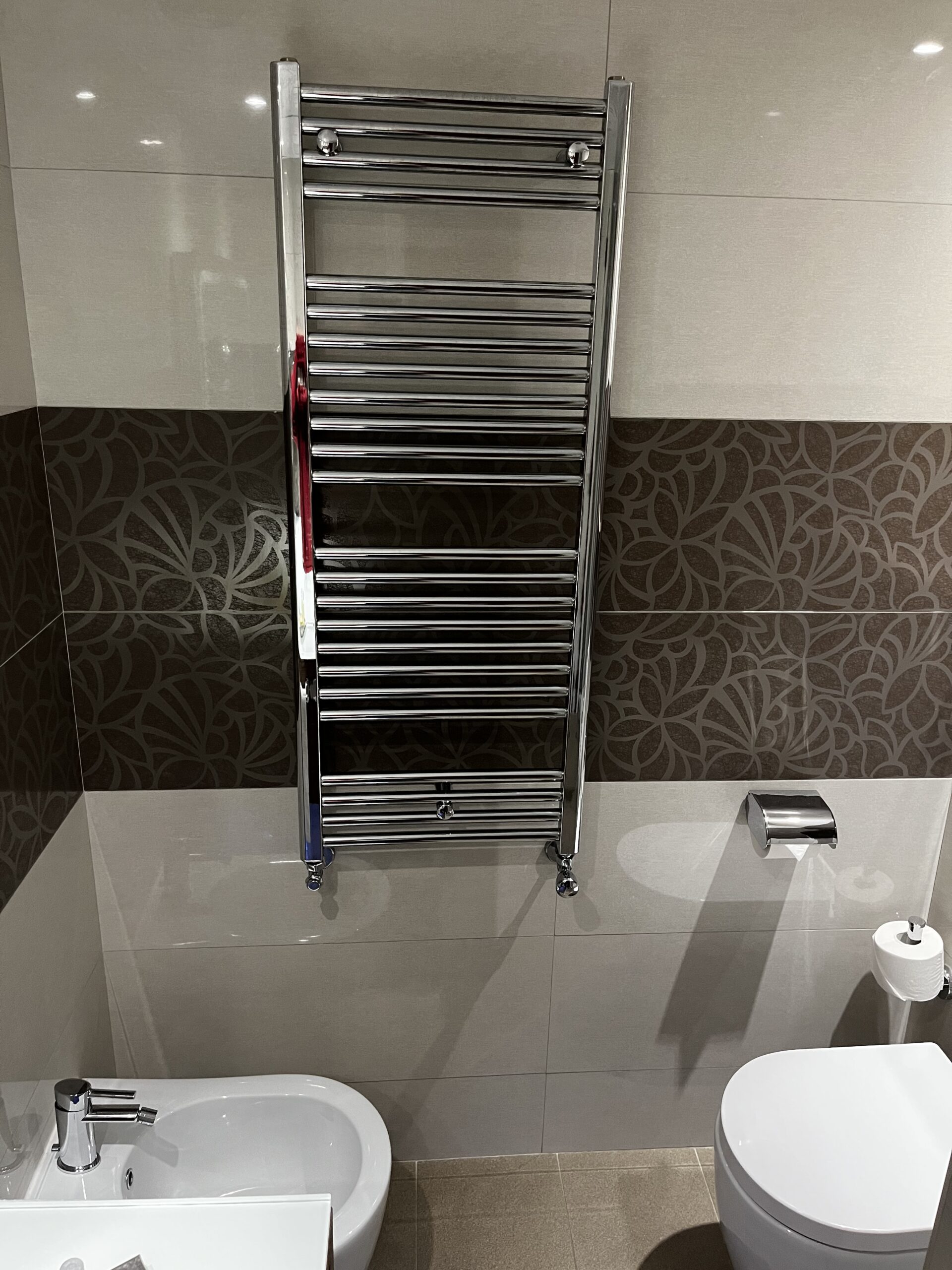 Chromener Handtuchheizkörper zwischen Bidet und WC an gefliester Badezimmerwand.