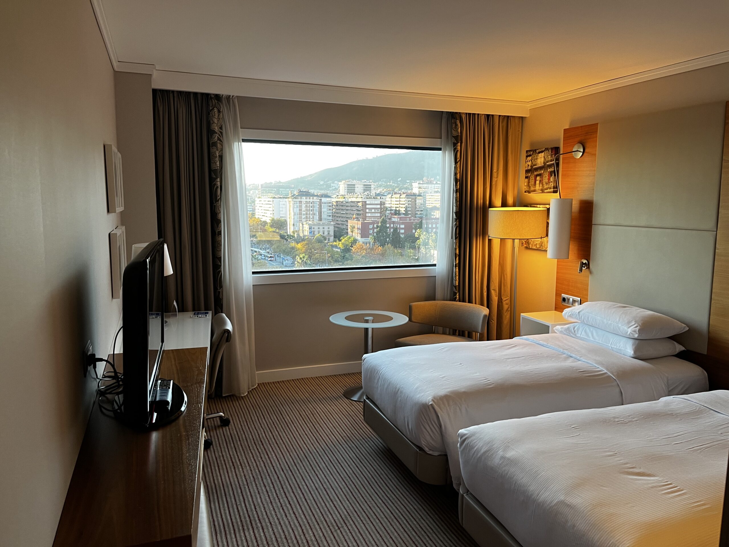 Hotelzimmer mit zwei Einzelbetten, TV, Sessel und großem Fenster mit Stadtblick auf Barcelona.