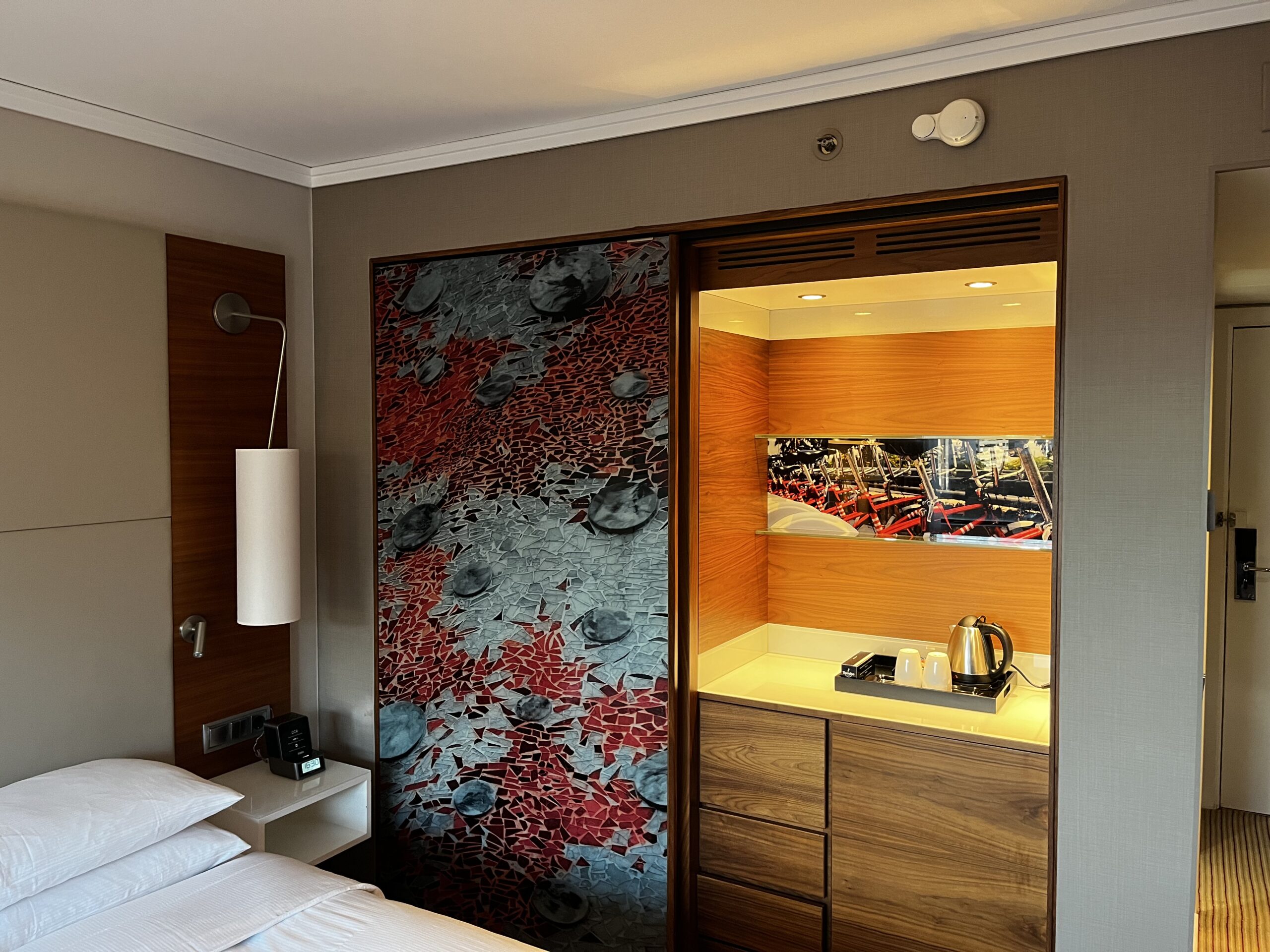 Hotelzimmer mit Bett, Wandleuchte, dekorativem Mosaikschiebeelement und beleuchtetem Minibar-Einbauschrank mit Wasserkocher.