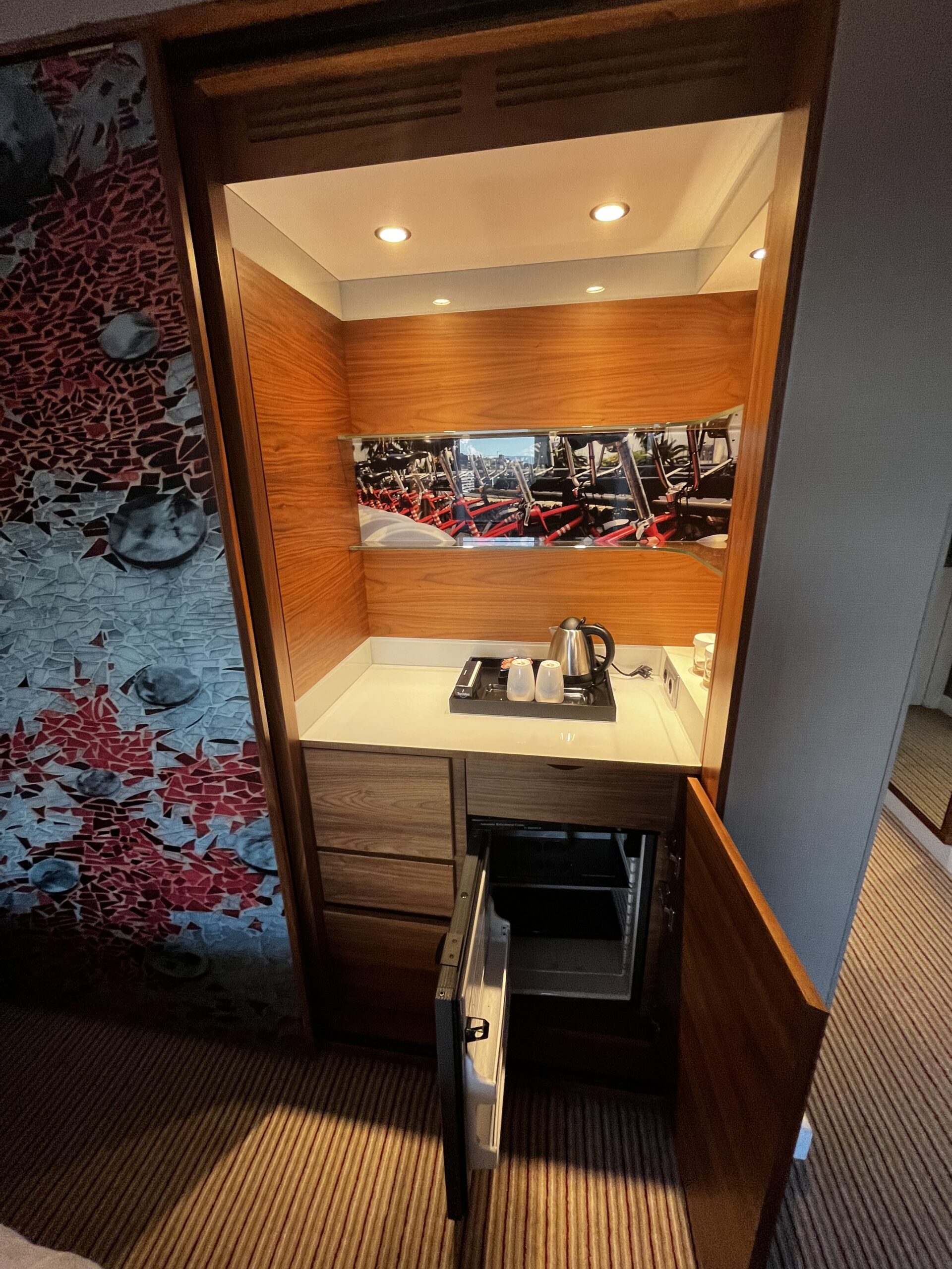 Minibar-Nische im Hotelzimmer mit Wasserkocher, Tassen und geöffnetem Kühlschrank, Holzverkleidung.