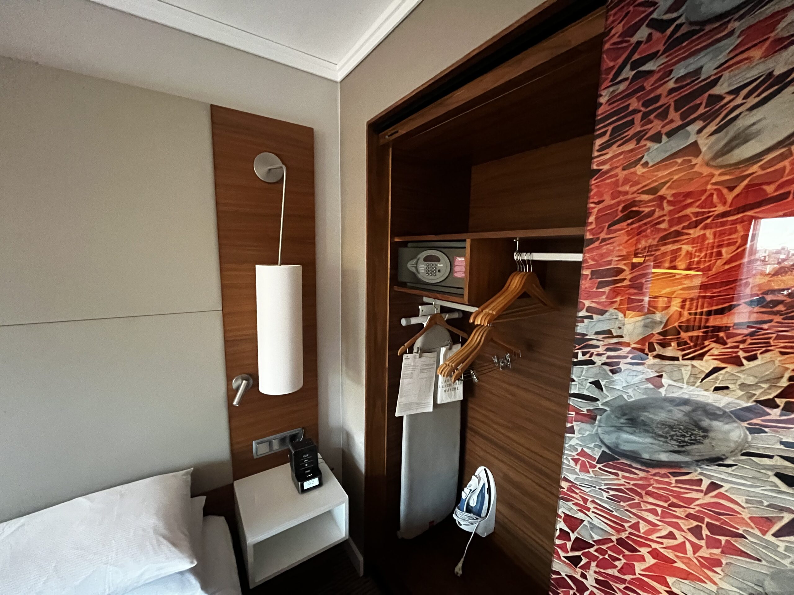 Hotelzimmer mit geöffnetem Kleiderschrank, Bügeleisen, Bügelbrett, Holzbügeln und Safe.