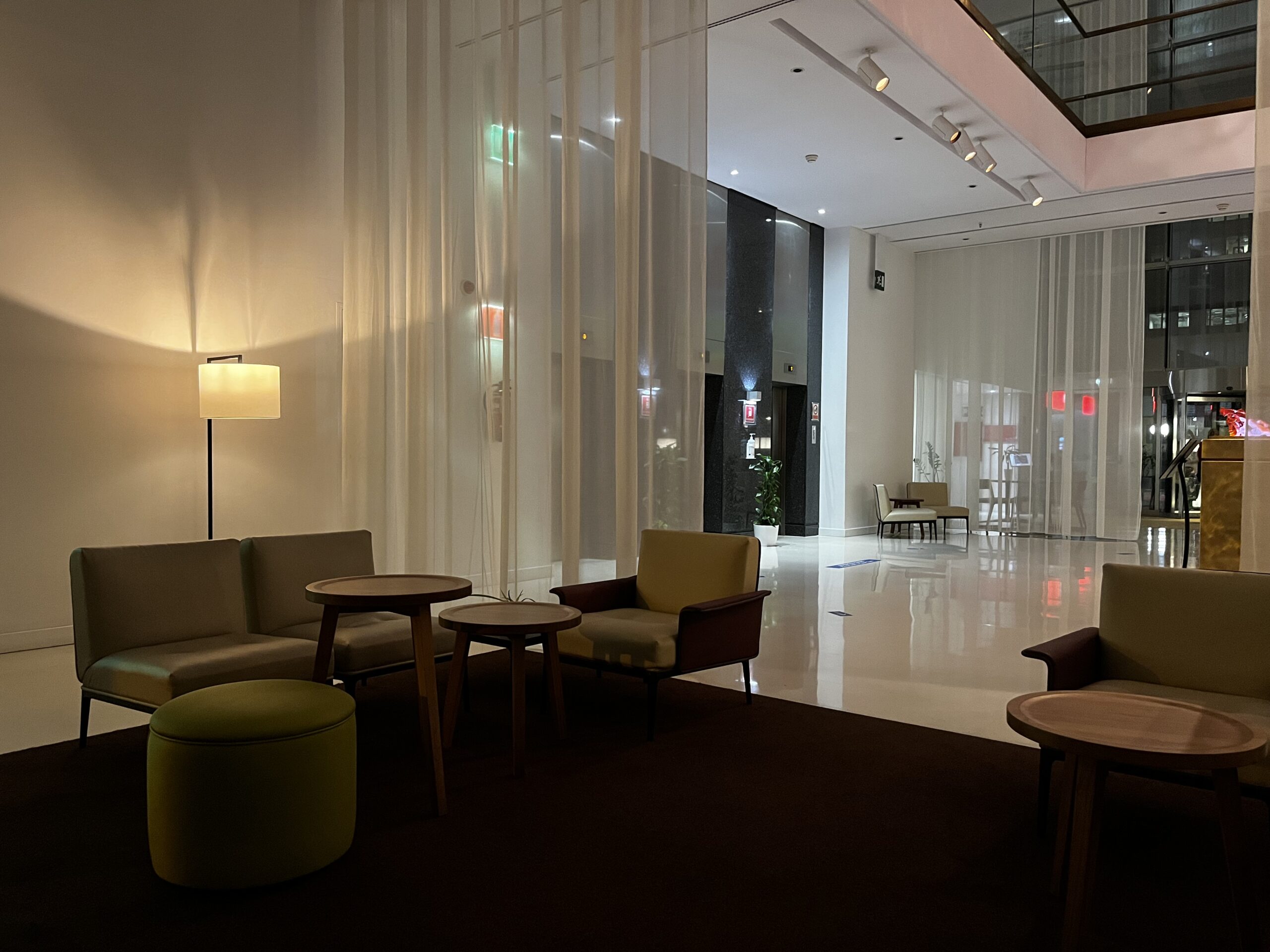 Hotellobby mit Sitzgruppen, Stehlampe, weißen Vorhängen und Blick auf den Eingangsbereich.