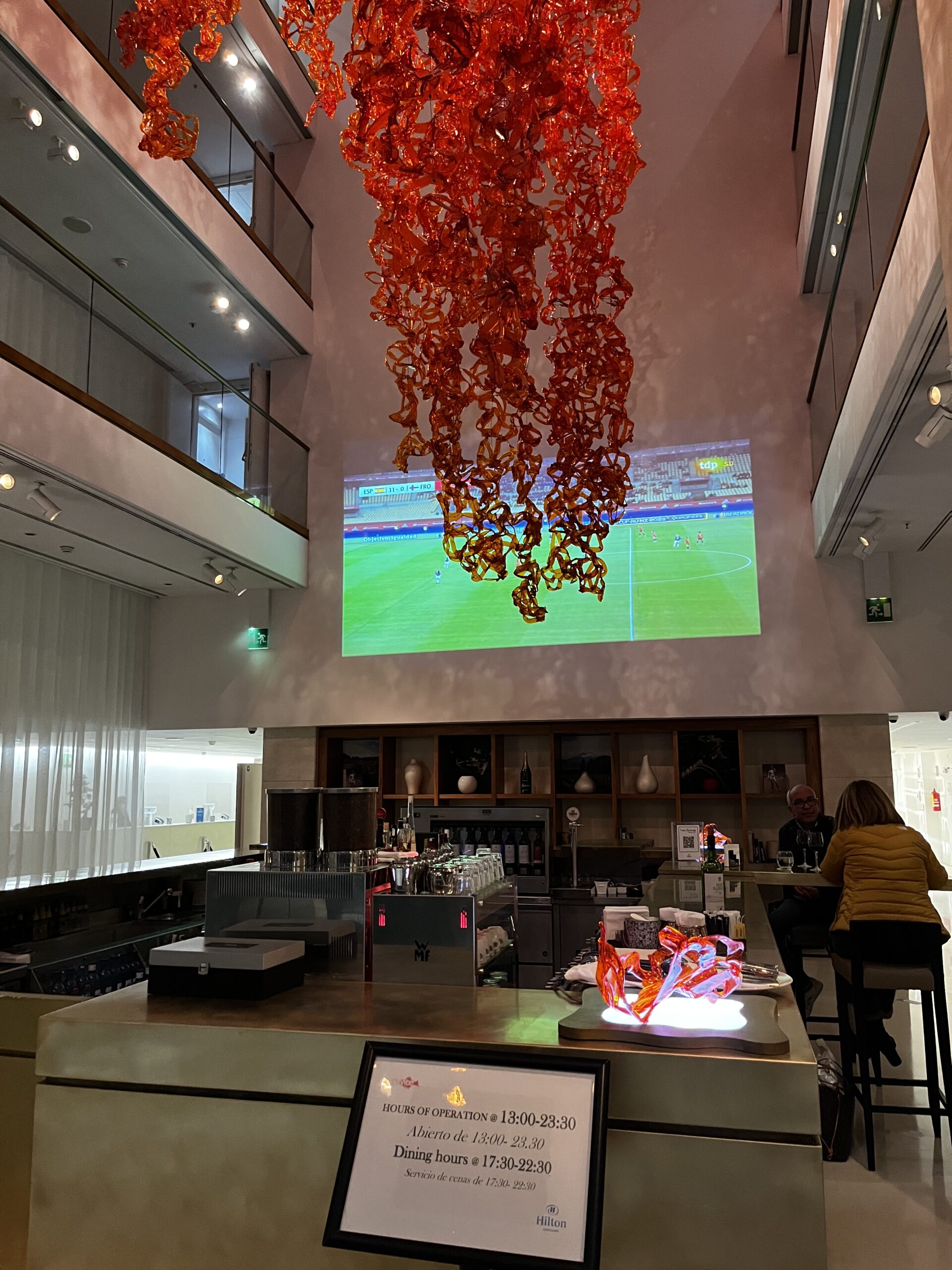Hotelbar mit roter Glaskunst-Deckeninstallation, Projektion eines Fußballspiels an der Wand, Öffnungszeiten-Schild im Vorderg