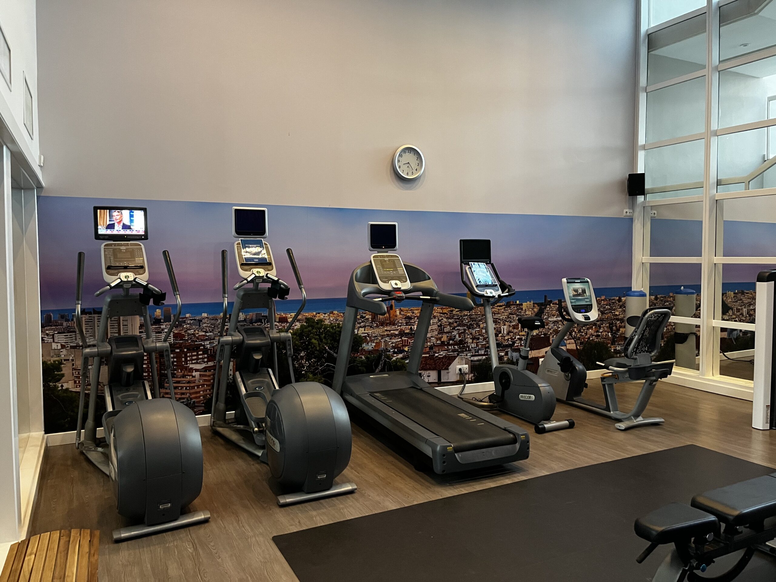 Fitnessraum mit zwei Crosstrainern, einem Laufband und zwei Fahrradergometern vor einem Stadtpanorama-Wandbild.