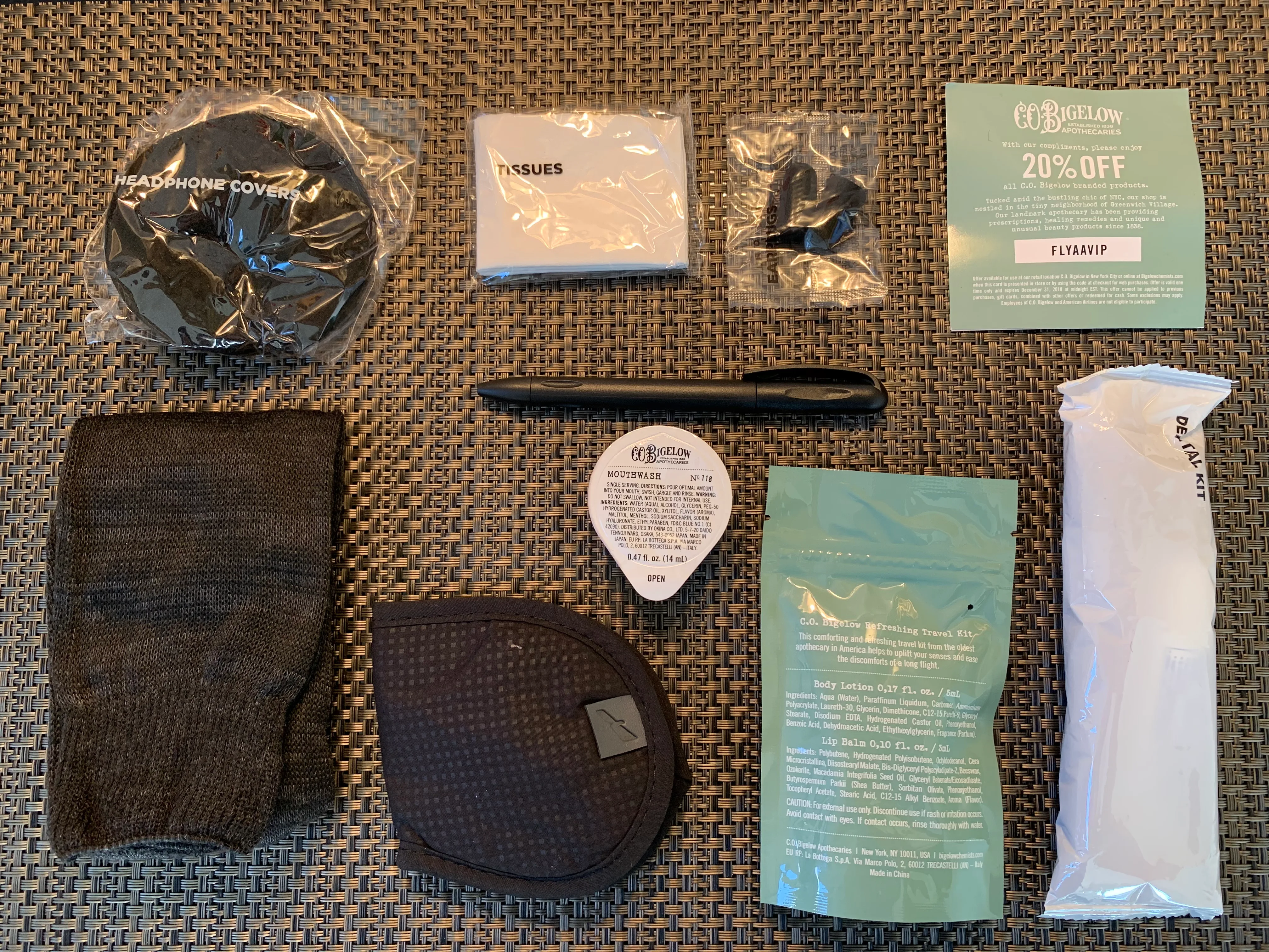 Inhalt des American Airlines Business Class Amenity Kits flach ausgebreitet: Kopfhörerhülle, Taschentücher, Zahnbürsten-Set, C.O. Bigelow Mundspülung und Bodylotion, Schlafmaske, Kugelschreiber, Lippenbalsam und FlyAndVIP 20 %-Gutschein