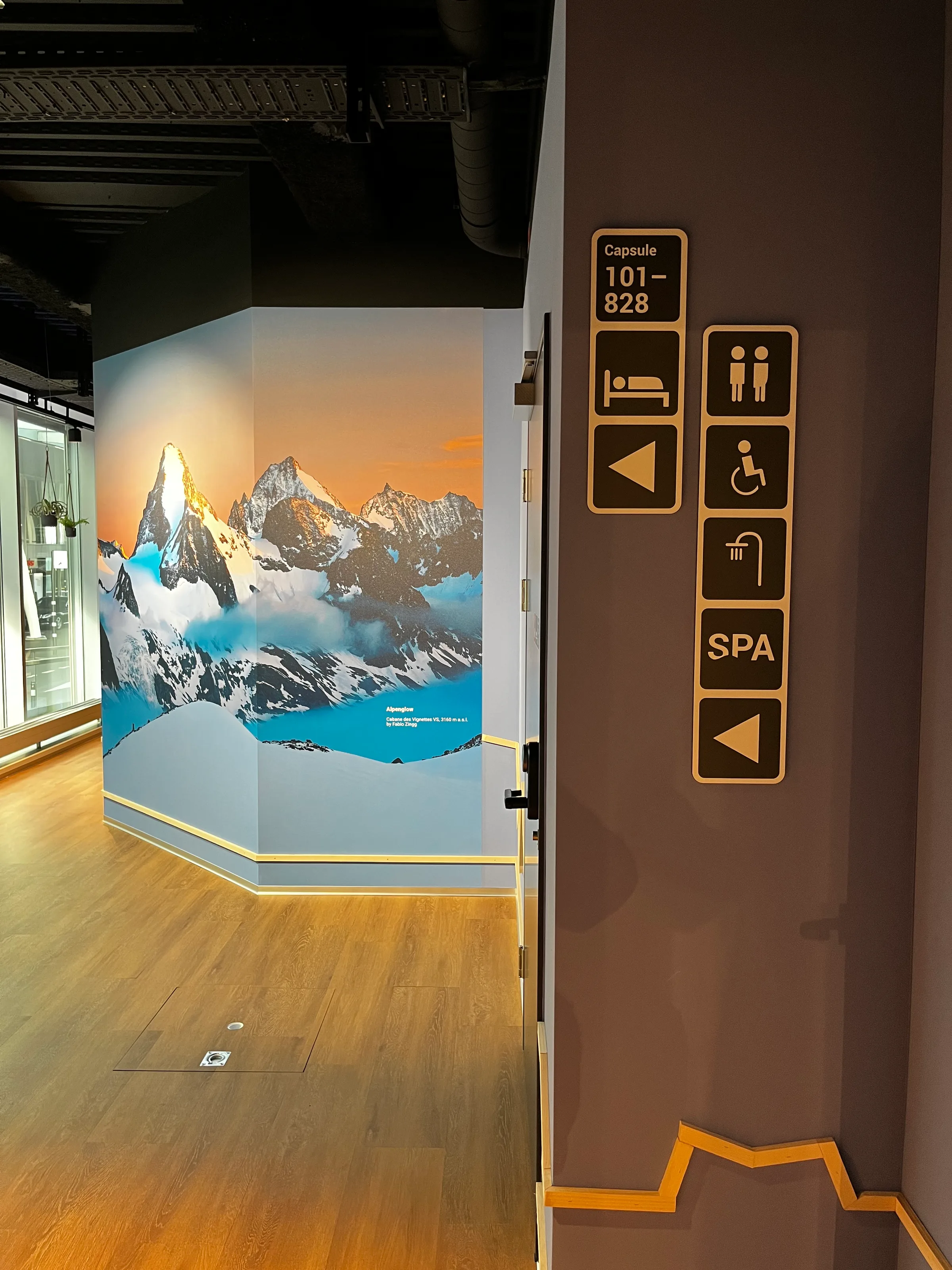 Gebäudeinnenraum mit Bergmotiv an der Wand und Hinweisschildern für Kapselfahrzeuge und Badezimmer
