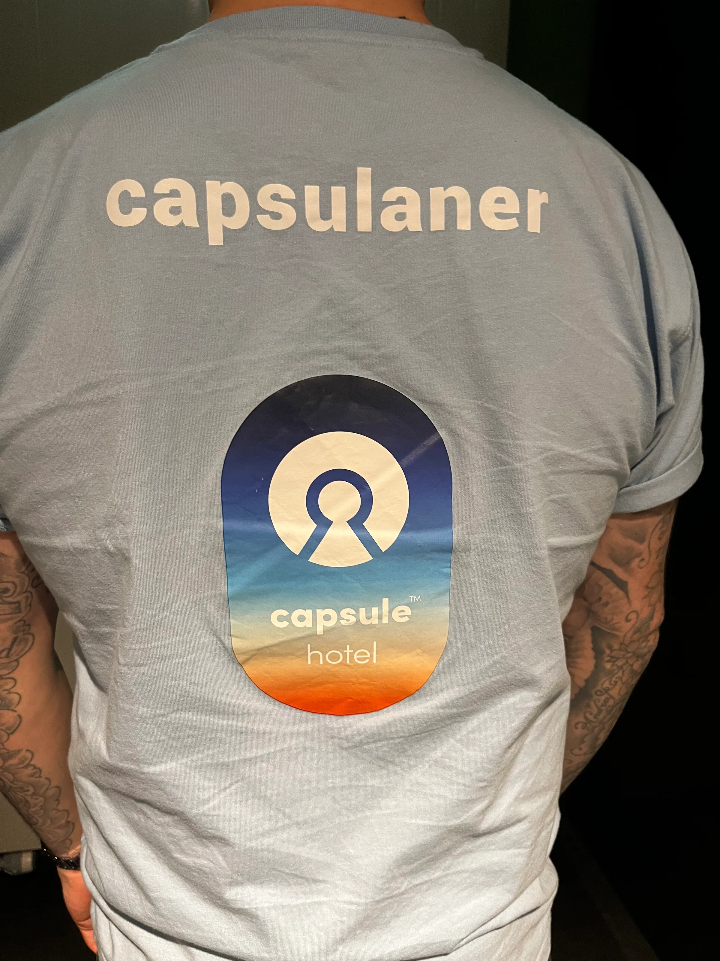 Rückansicht eines T-Shirts mit Aufdruck 'capsulaner' und Logo 'capsule hotel'