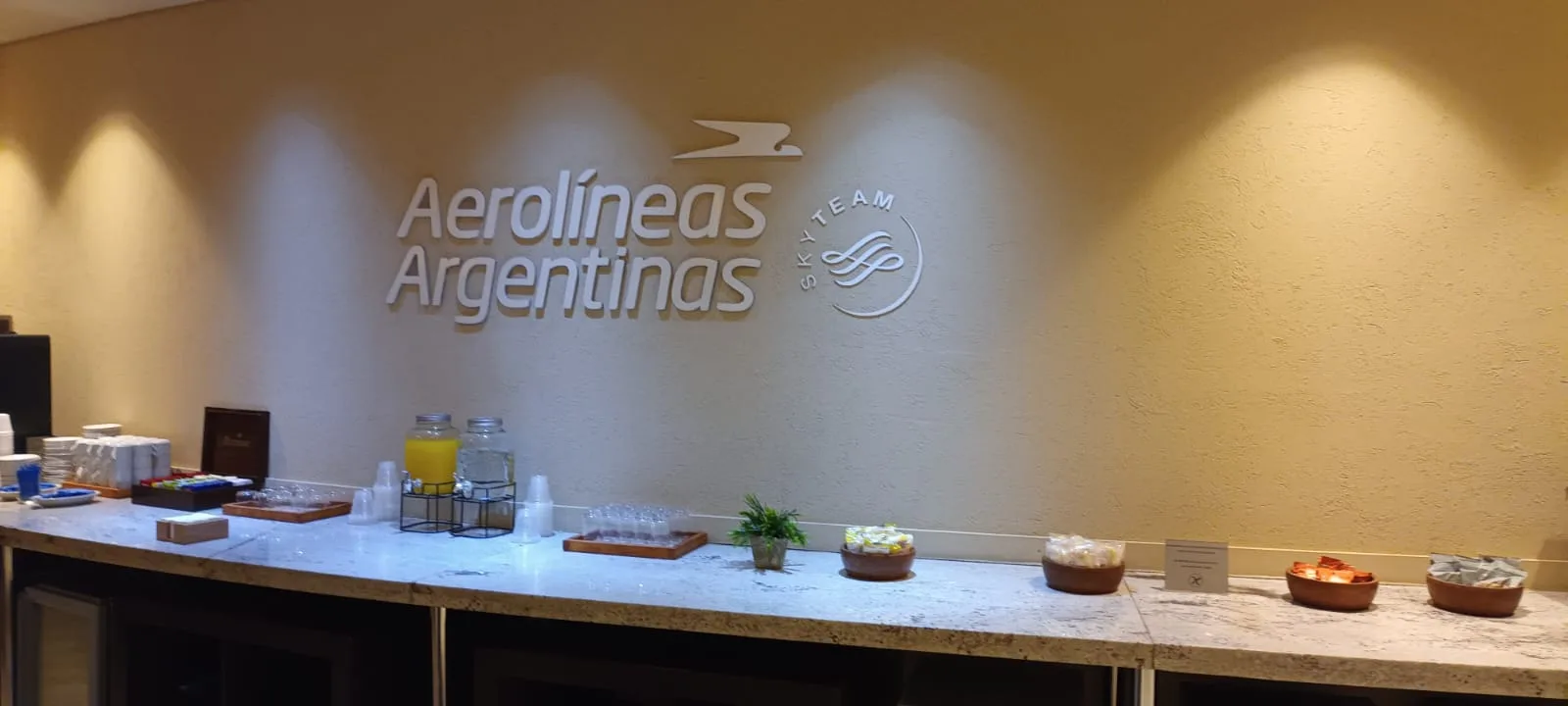 Lounge-Tresen mit Getränken und Snacks vor Wand mit Aerolíneas Argentinas-Logo