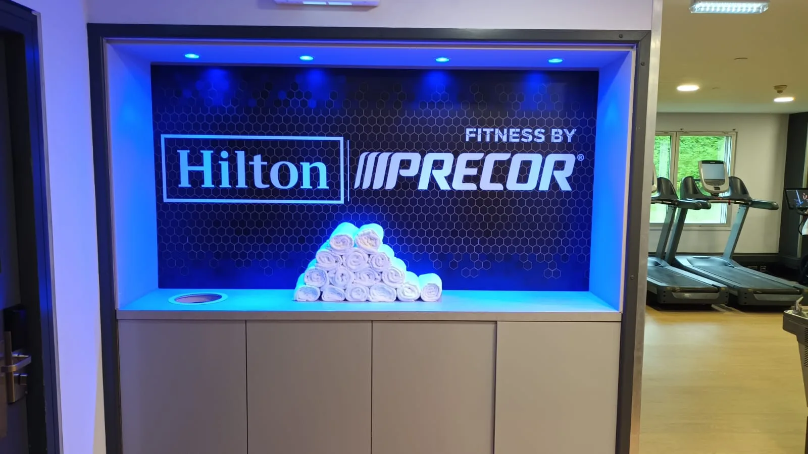 Fitnessbereich mit Handtuchpyramide und Logos von Hilton und Precor