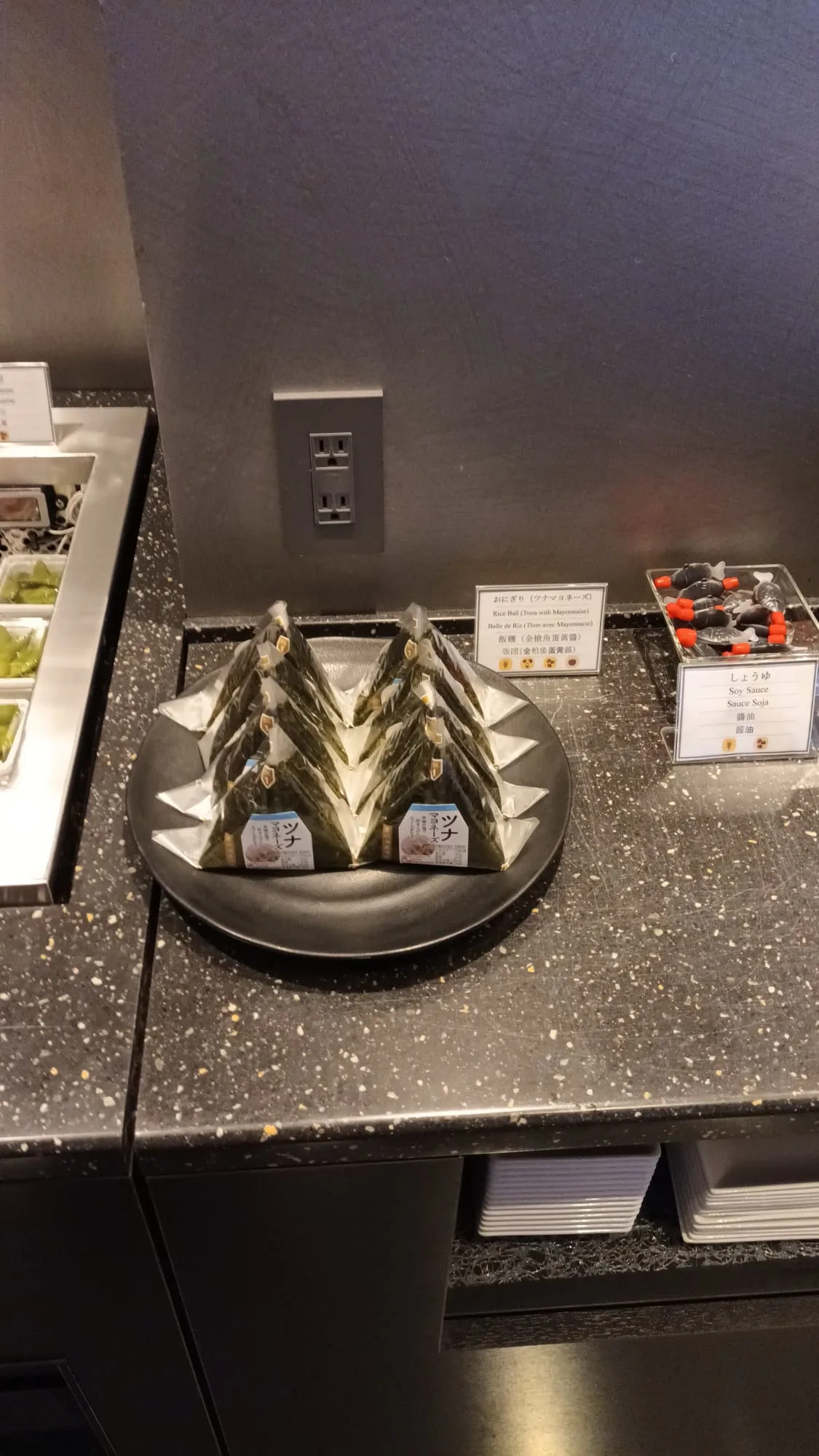 Drei aufgerollte Onigiri auf einem schwarzen Teller mit Essstäbchen und Sojasauce.