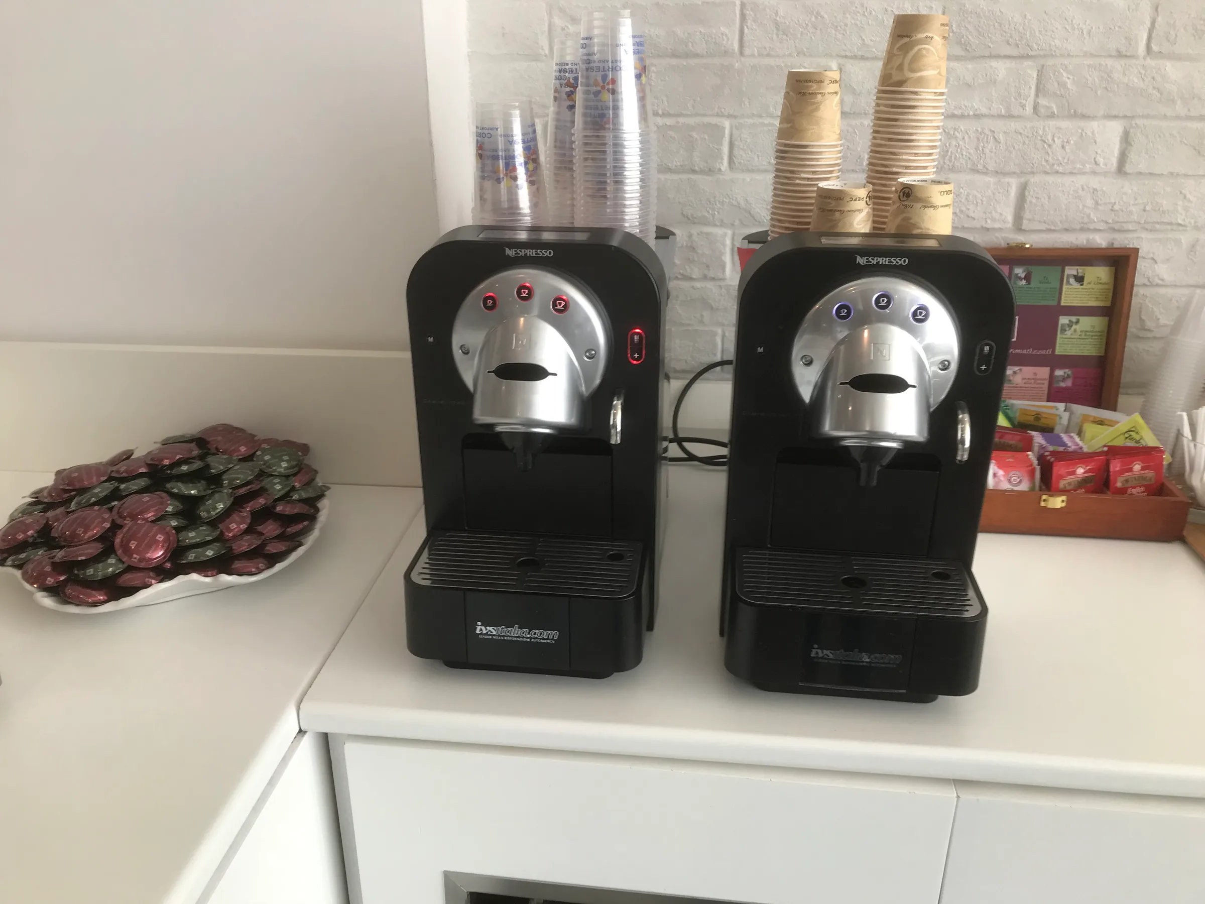 Zwei Nespresso-Kaffeevollautomaten auf weißem Sideboard mit Kaffeepads und Bechern