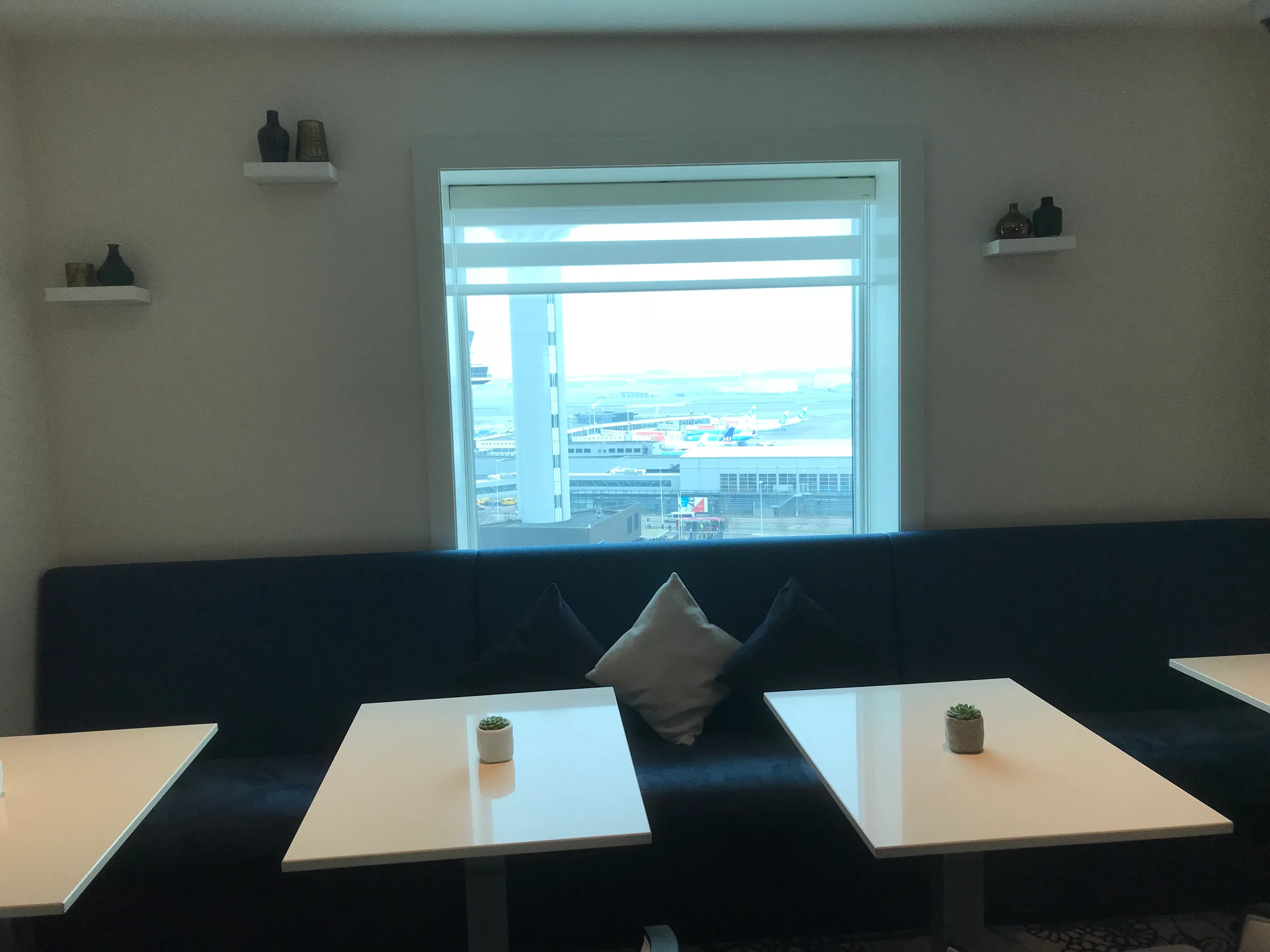 Hotelzimmer mit Sitzbank und Tischen vor Fenster mit Flughafenblick