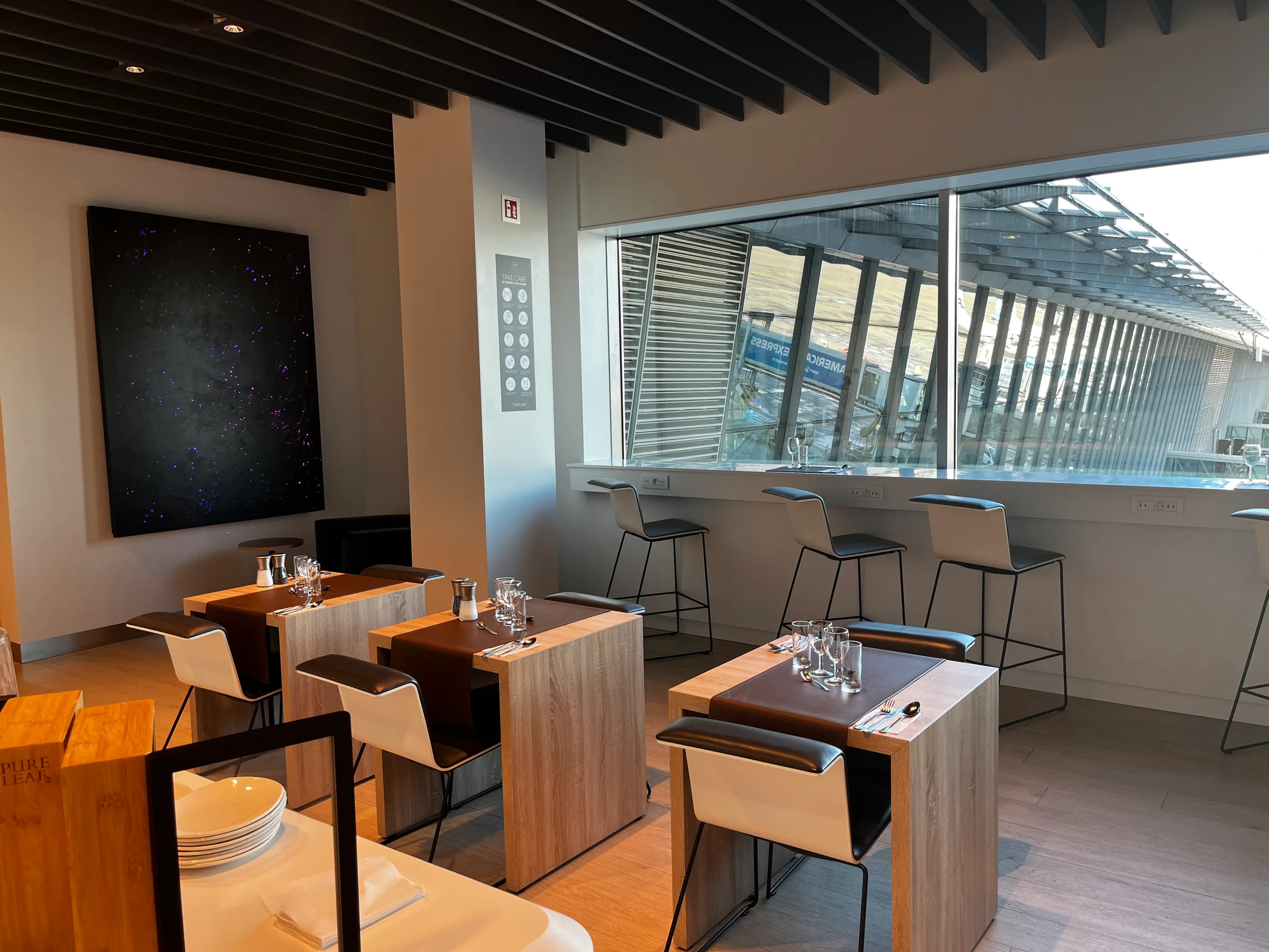 Lounge mit Holztischen und Stühlen, Fensterfront mit Blick auf Flughafen